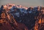 Veduta dalla catena delle Tre Croci (piccole Dolomiti) verso la prima luce del giorno, che illumina il  Monte Pasubio. Visibili in basso il Sacrario militare e in alto, tra la strada degli eroi a sin e la strada dell 52 gallerie a destra, il Rifugio Papa. ancora nell'ombra. Gennaio 2024